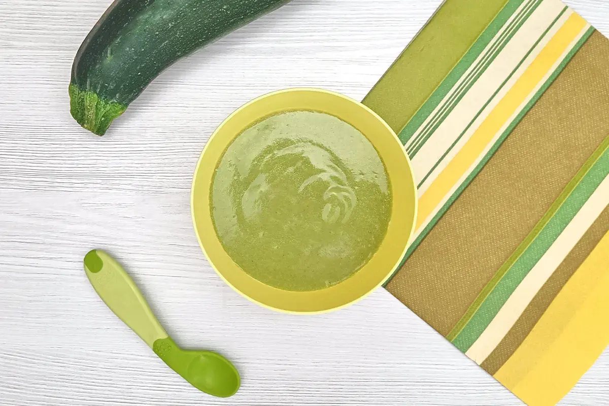 Un plato con puré de calabacín bebé, una cuchara verde y un calabacín entero sobre una mesa de madera.