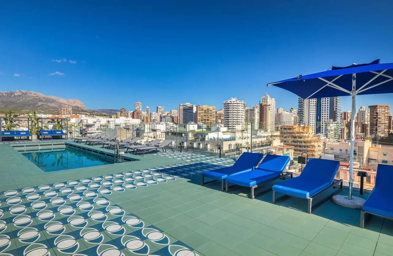 Terraza con piscina y tumbonas azules en Barceló Benidorm, con vistas a la ciudad y montañas.