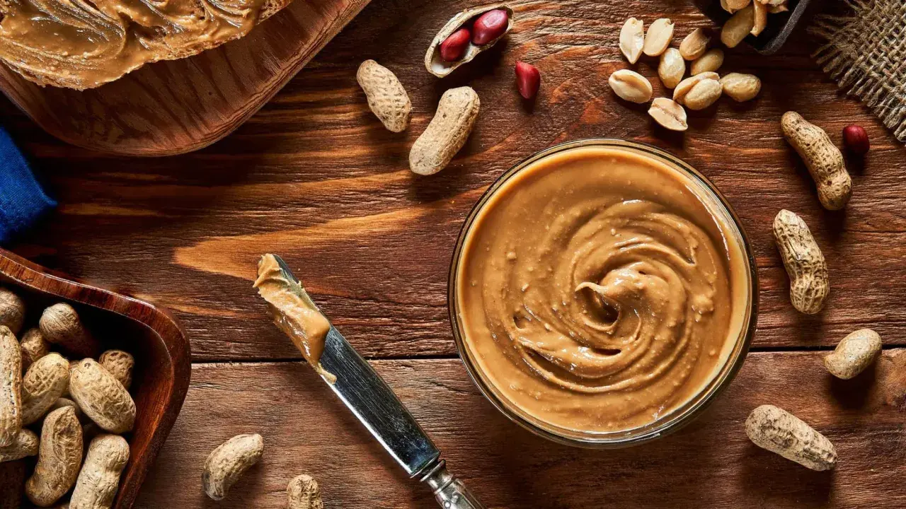 Cremige Erdnussbutter in einer Schüssel, daneben Erdnüsse und ein Messer. Erdnussbutter gesund und lecker!