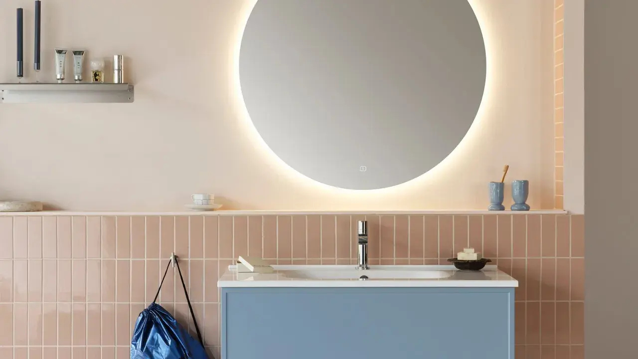 Ett modernt badrum med en stor, upplyst rund spegel som ger en känsla av rymd. Att inreda med speglar är smart! Blått kommod och rosa kakel skapar en mjuk atmosfär.