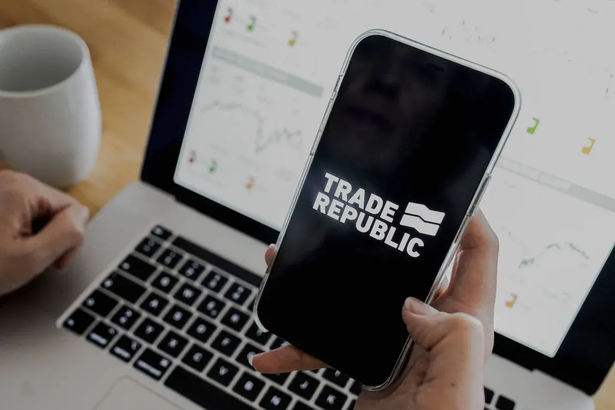Person prüft den Trade Republic Quartalsbericht auf dem Smartphone, während der Laptop mit Finanzdiagrammen im Hintergrund läuft.