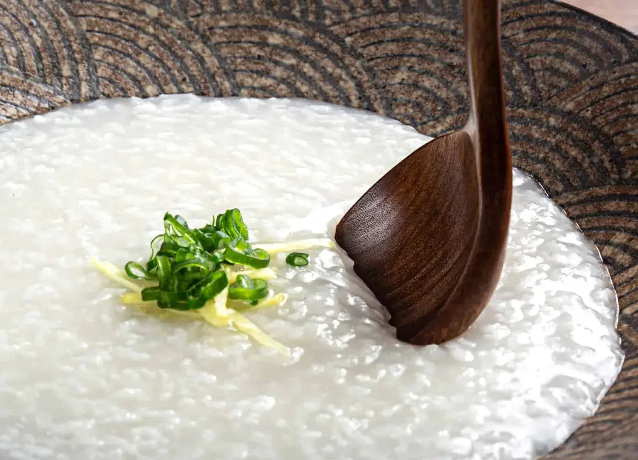 A comforting bowl of Japanese rice porridge (okayu) garnished with fresh scallions and ginger, ready to be enjoyed with a wooden spoon.