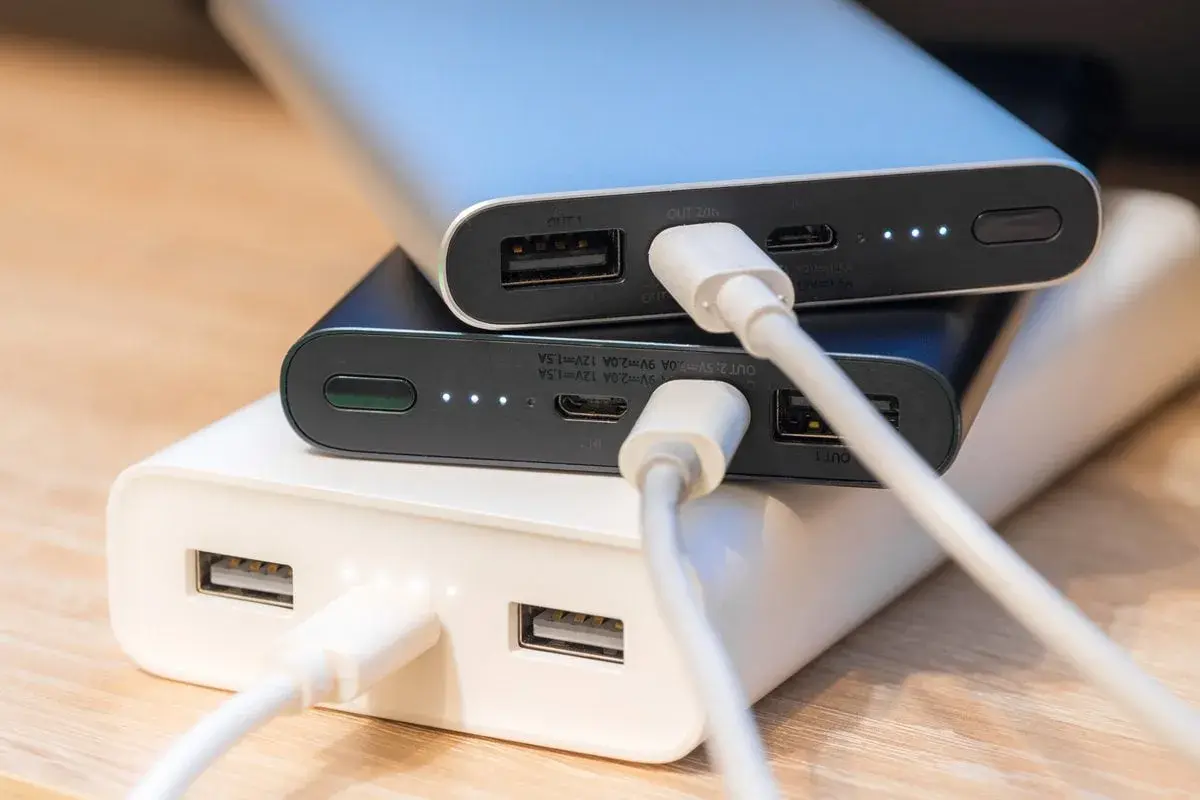 Trzy powerbanki w kolorach białym, czarnym i srebrnym, podłączone kablami USB. To przenośne ładowarki, które pomogą naładować urządzenia w podróży.