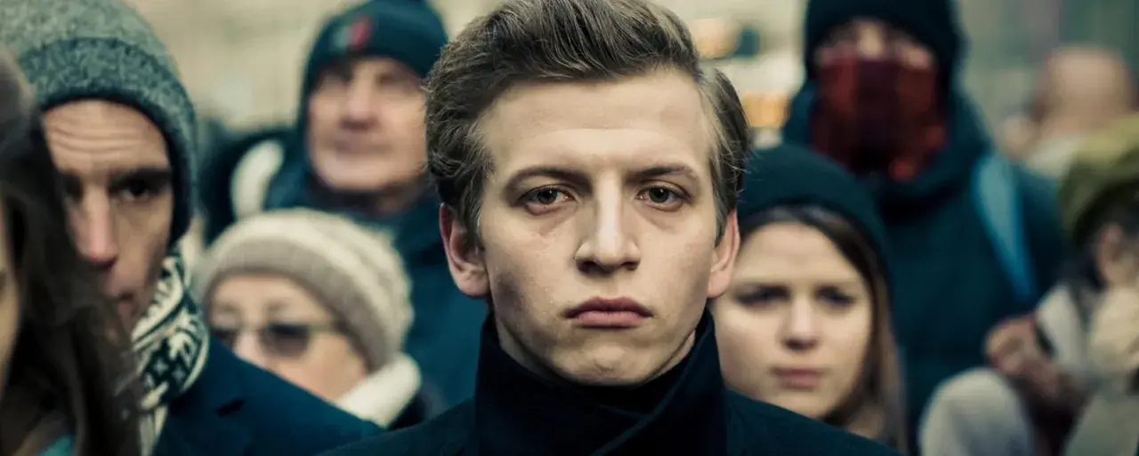 Młody mężczyzna na pierwszym planie, otoczony tłumem. Jego wyraz twarzy sugeruje głębokie przemyślenia, jakby przygotowywał się do roli w jednym z filmów Macieja Musiałowskiego.