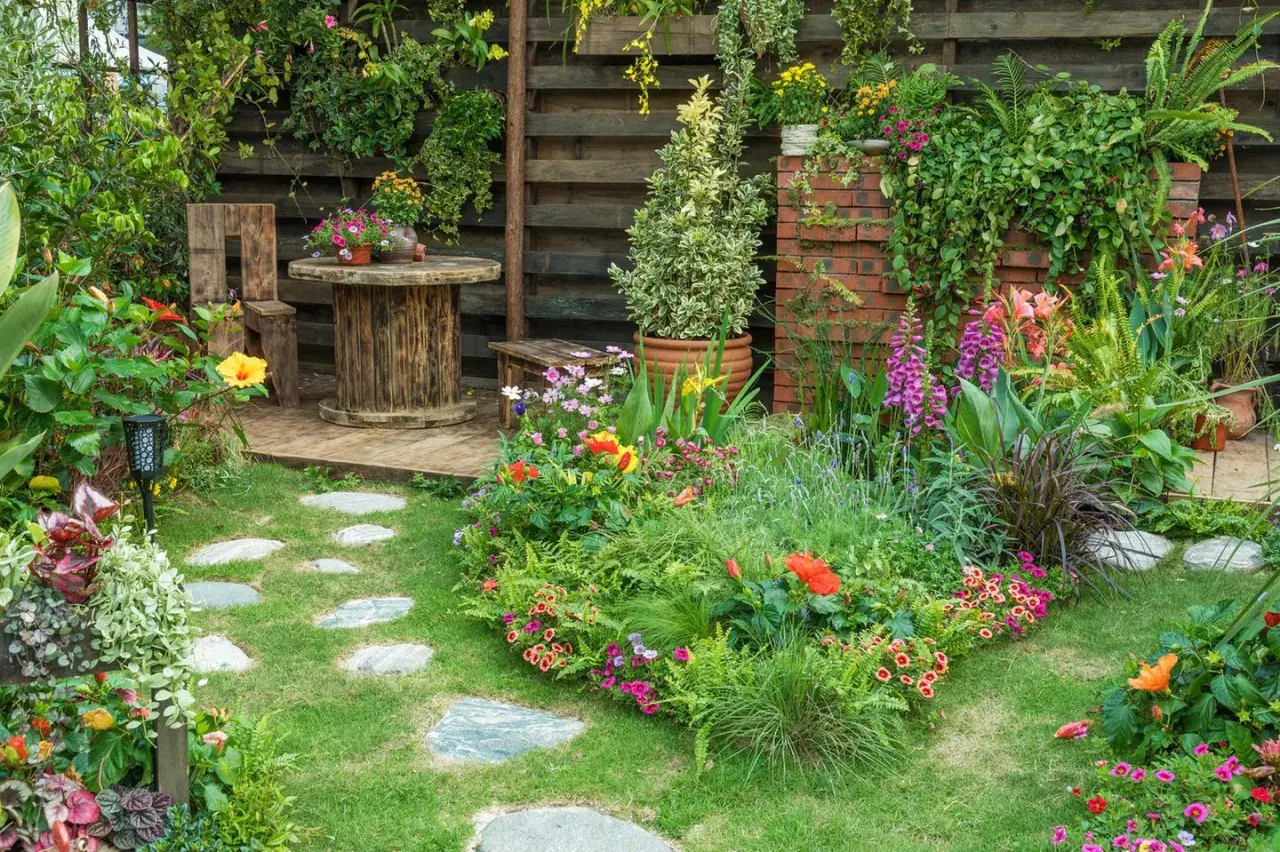 Ein üppiger, naturnaher Garten mit bunten Blumen, grünen Pflanzen und einem rustikalen Sitzbereich. Steinplatten führen durch das satte Grün.