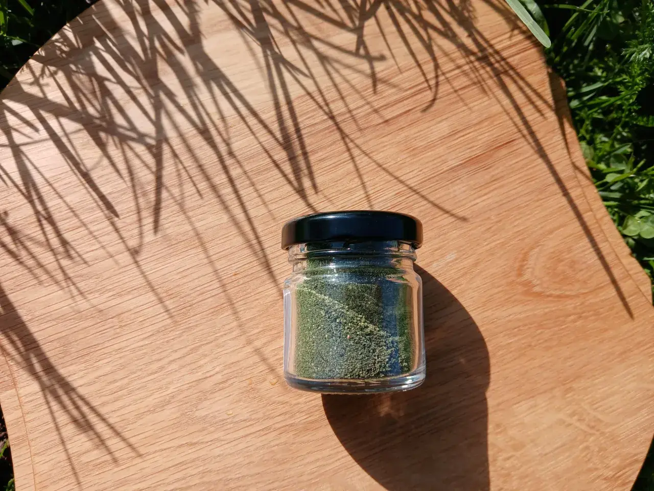 Un piccolo barattolo di vetro contiene polvere di ortica verde brillante, posato su un tagliere di legno con ombre di foglie.