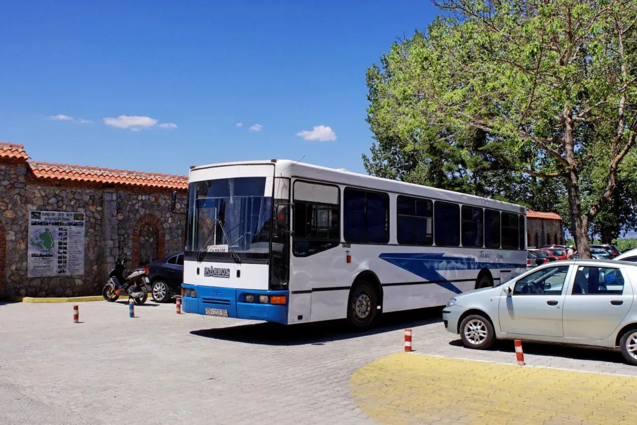 Biały macedoński autobus z niebieskimi pasami stoi na parkingu obok kamiennego muru i drzew.