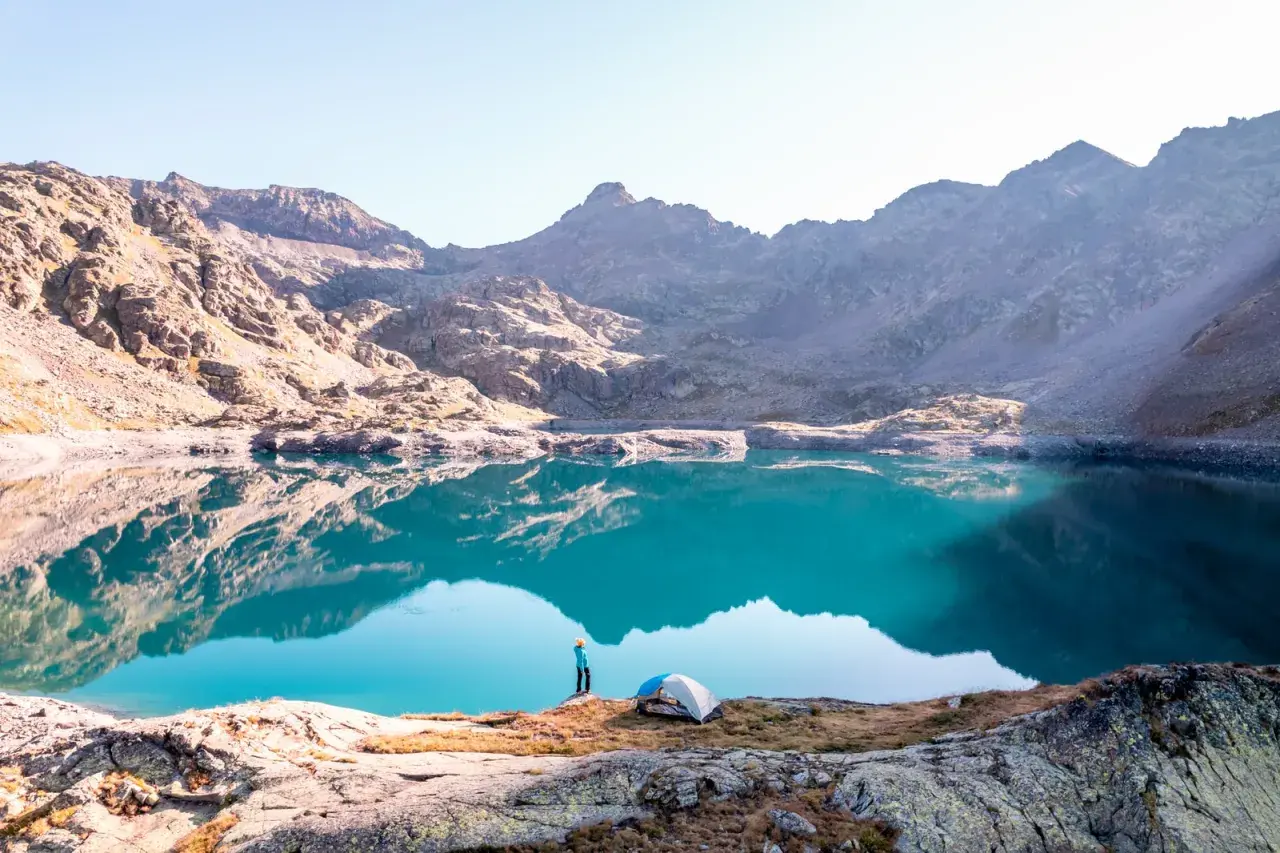 Un randonneur admire un lac turquoise aux reflets parfaits dans le **parc du Mercantour**. Une tente est installée sur la rive rocheuse.