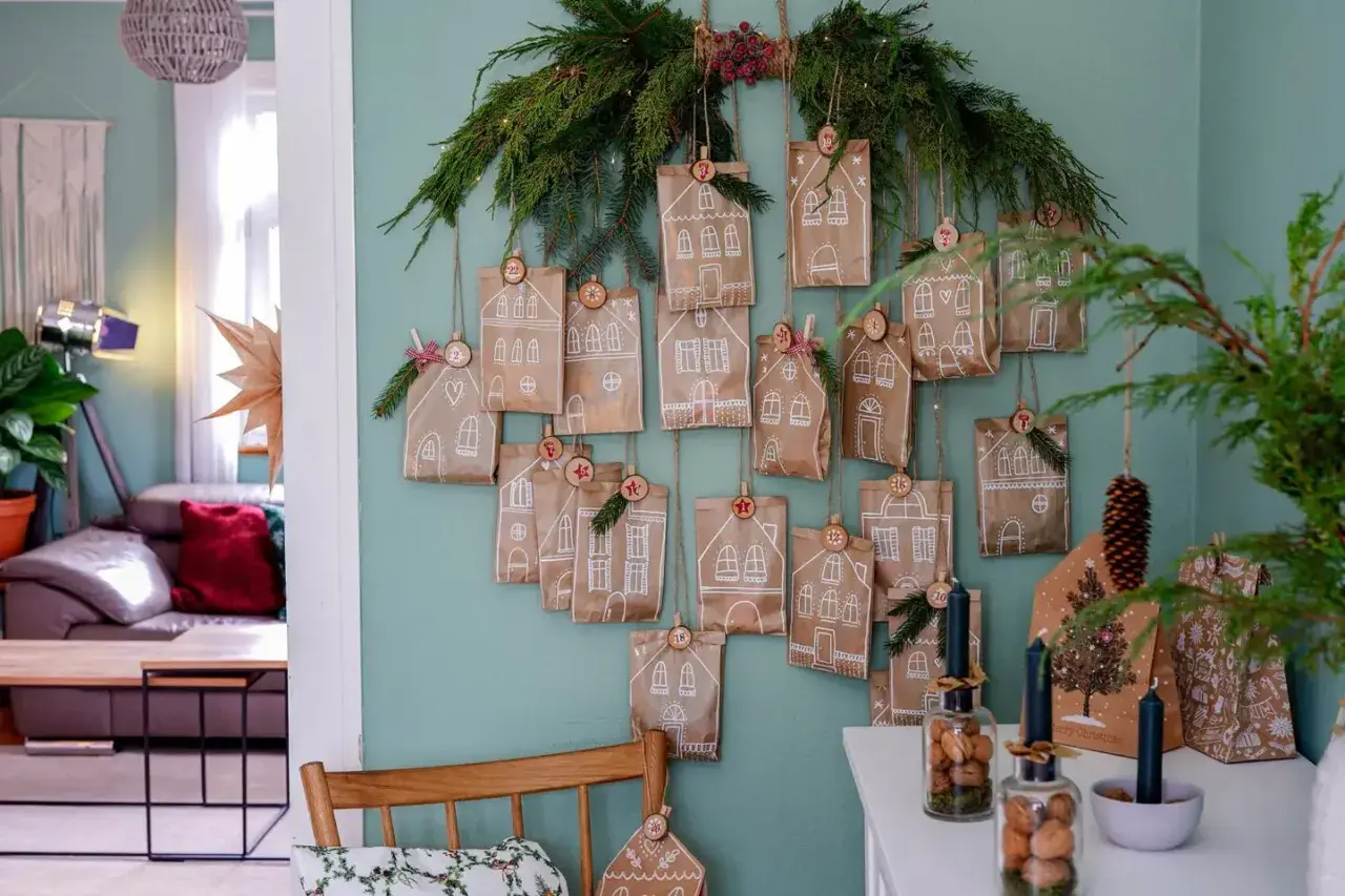 Selbstgemachter Adventskalender für Erwachsene: Kleine Papiertüten mit Hausmotiven und Zahlen hängen an Tannenzweigen.