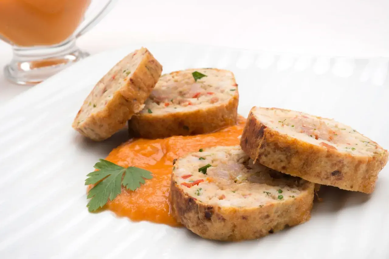 Rollo de bonito cortado en rodajas, servido sobre puré de pimientos y decorado con perejil.