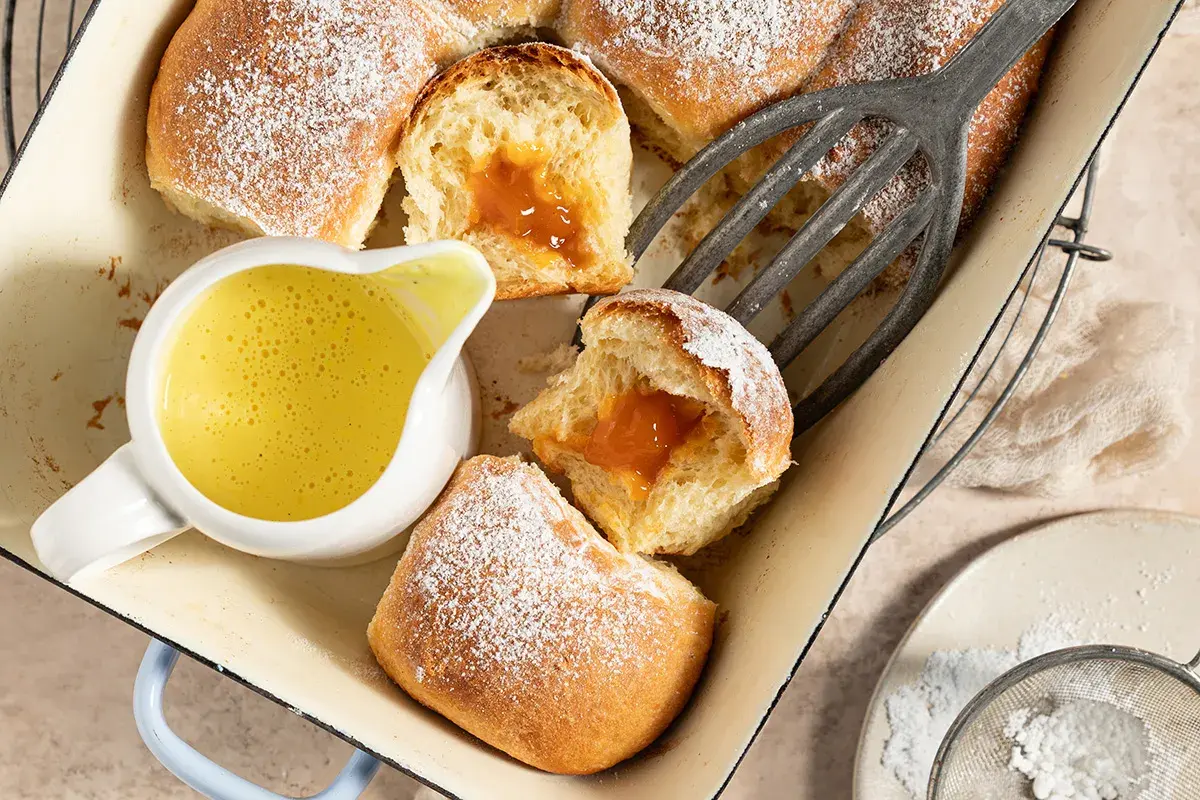 Gefüllte Buchteln mit Puderzucker und einer Schüssel cremiger Vanillesauce aus Puddingpulver.