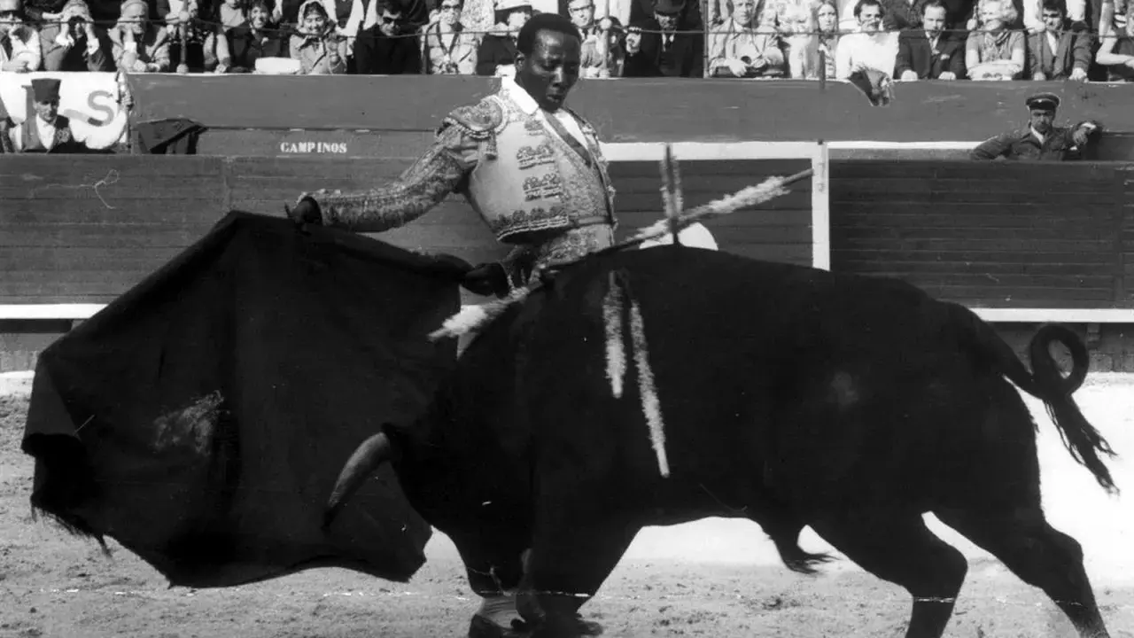 Un torero negro, con traje de luces, se enfrenta a un toro bravo en la arena. El público observa desde las gradas.