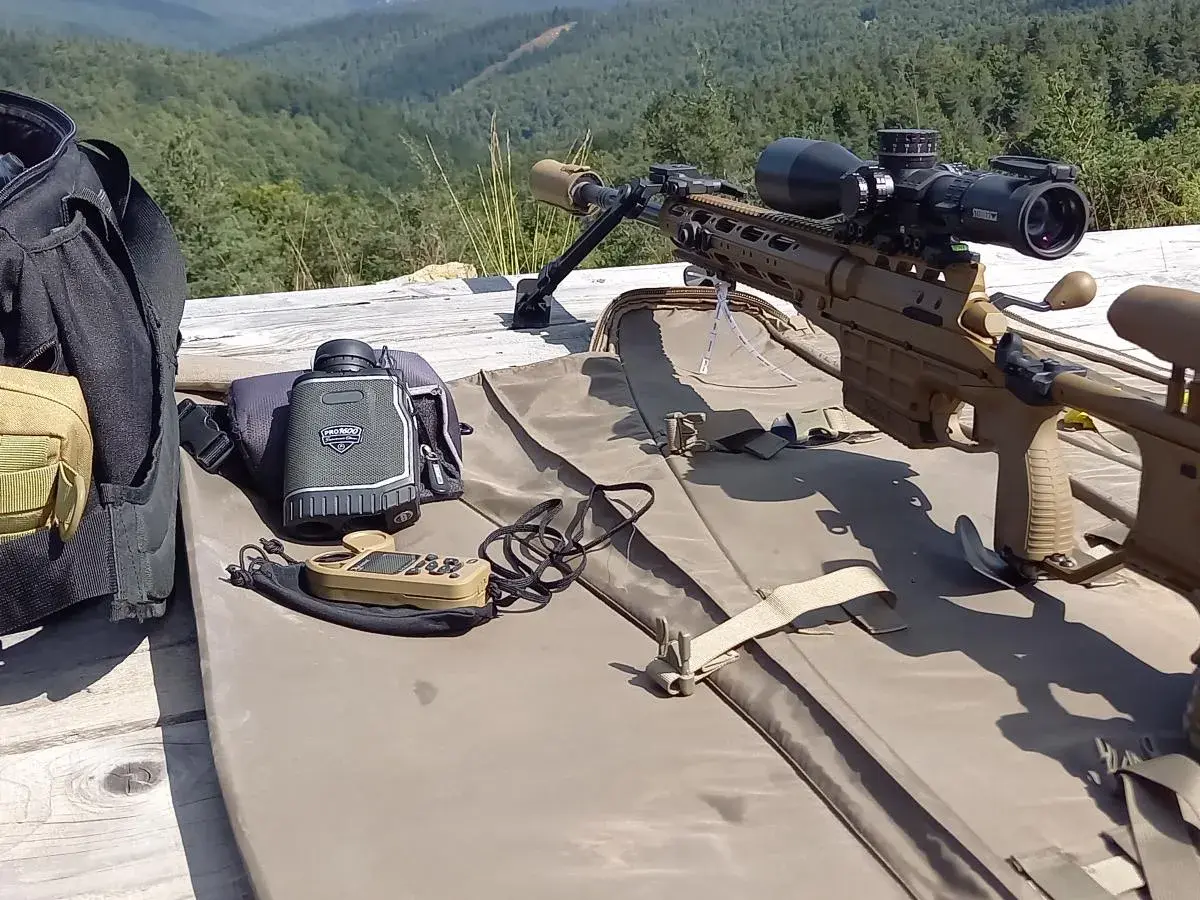 Carabine de précision beige, lunette, télémètre et sac de transport sur une table en bois, avec une forêt en arrière-plan.