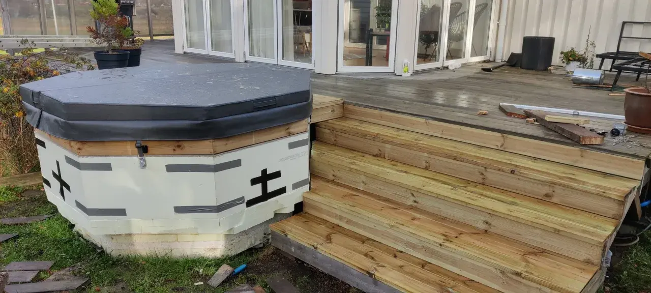 Pågår bygga badtunna. Isolerad stomme med trädetaljer och en grå presenning. Nya trätrappor leder upp till altanen.