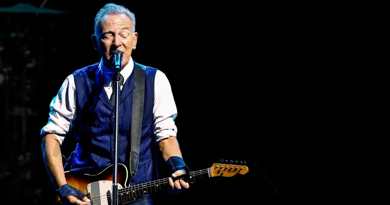 Bruce Springsteen z gitarą na scenie, w niebieskiej kamizelce i białej koszuli. Niezapomniane koncerty legendy rocka.