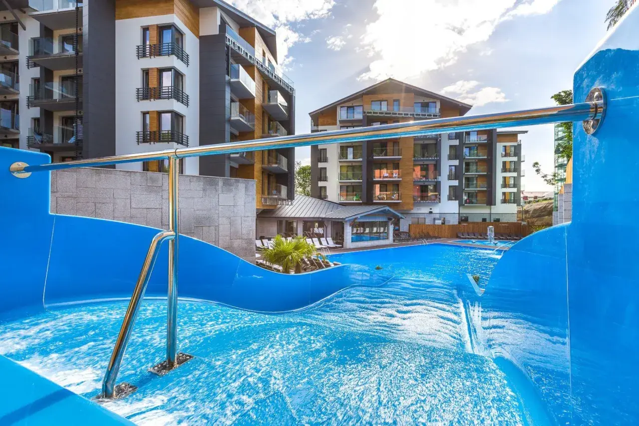 Basen z falą i zjeżdżalnią w hotelu Blue Mountain w Szklarskiej Porębie. Nowoczesne budynki hotelowe w tle.