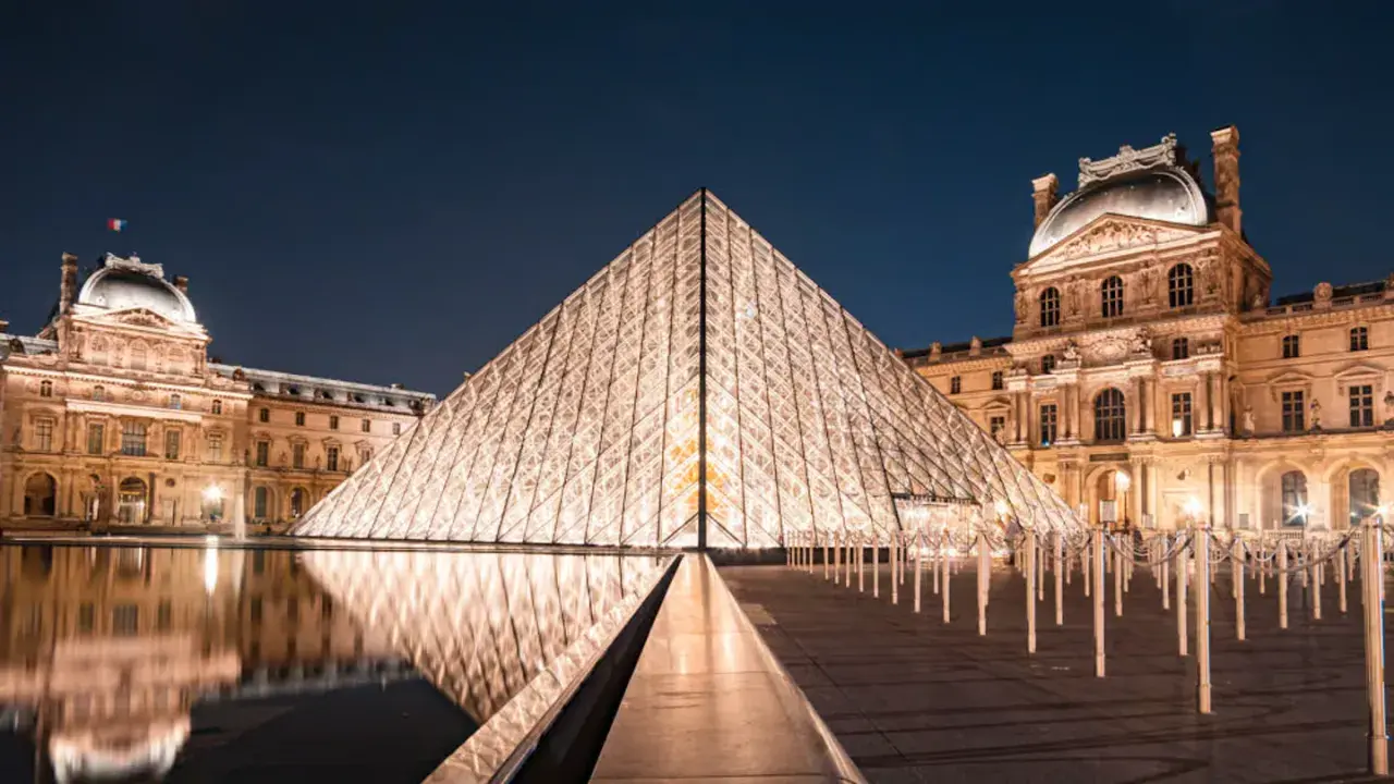 Piramide Luwru nocą z odbiciem w wodzie i oświetlonymi budynkami muzeum.