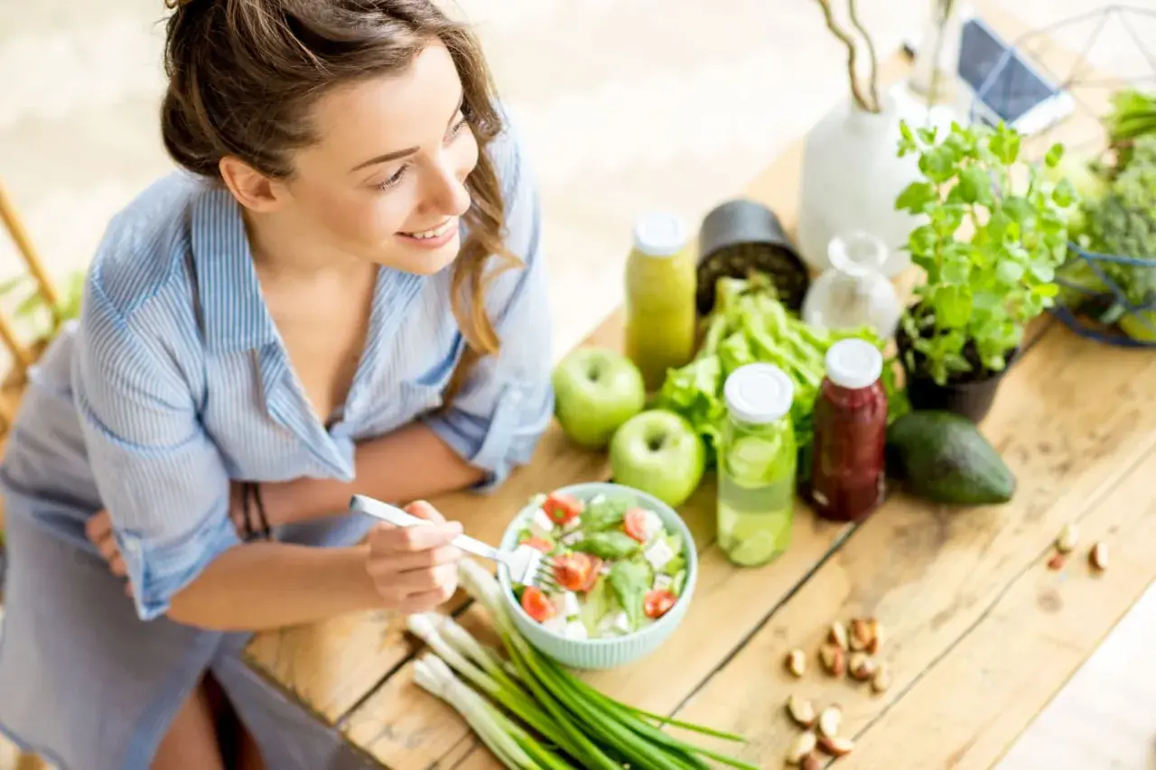 Junge Frau isst gesunden Salat. Viele gesunde Lebensmittel wie Obst, Gemüse und Nüsse sind auf dem Tisch, die reich an Cystein sind.