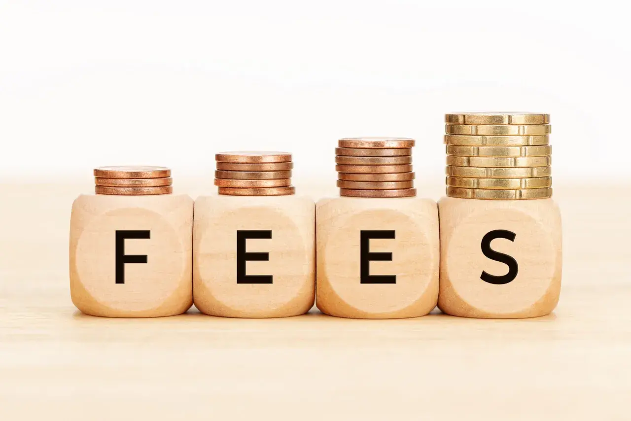 Cztery drewniane kostki z literami F, E, E, S tworzą napis FEES. Nad nimi piętrzą się monety, symbolizując rosnące opłaty.