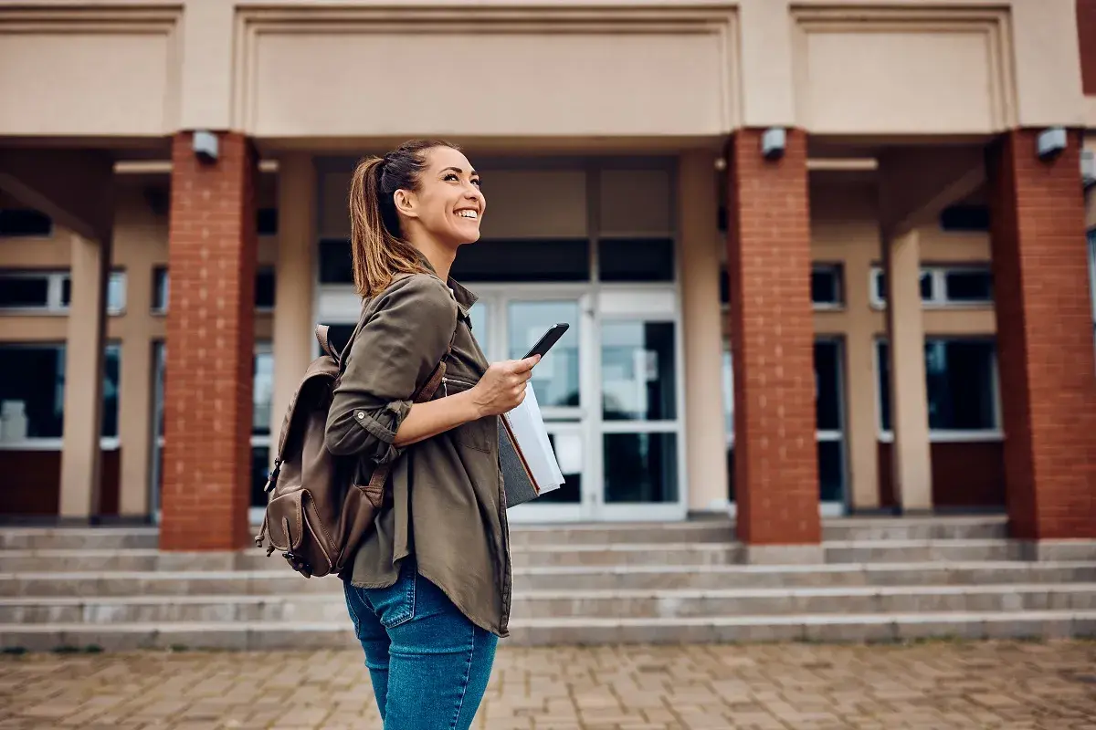 Uśmiechnięta studentka z plecakiem i telefonem przed budynkiem uczelni. Zastanawia się, jakie studia po rozszerzonym angielskim wybrać.