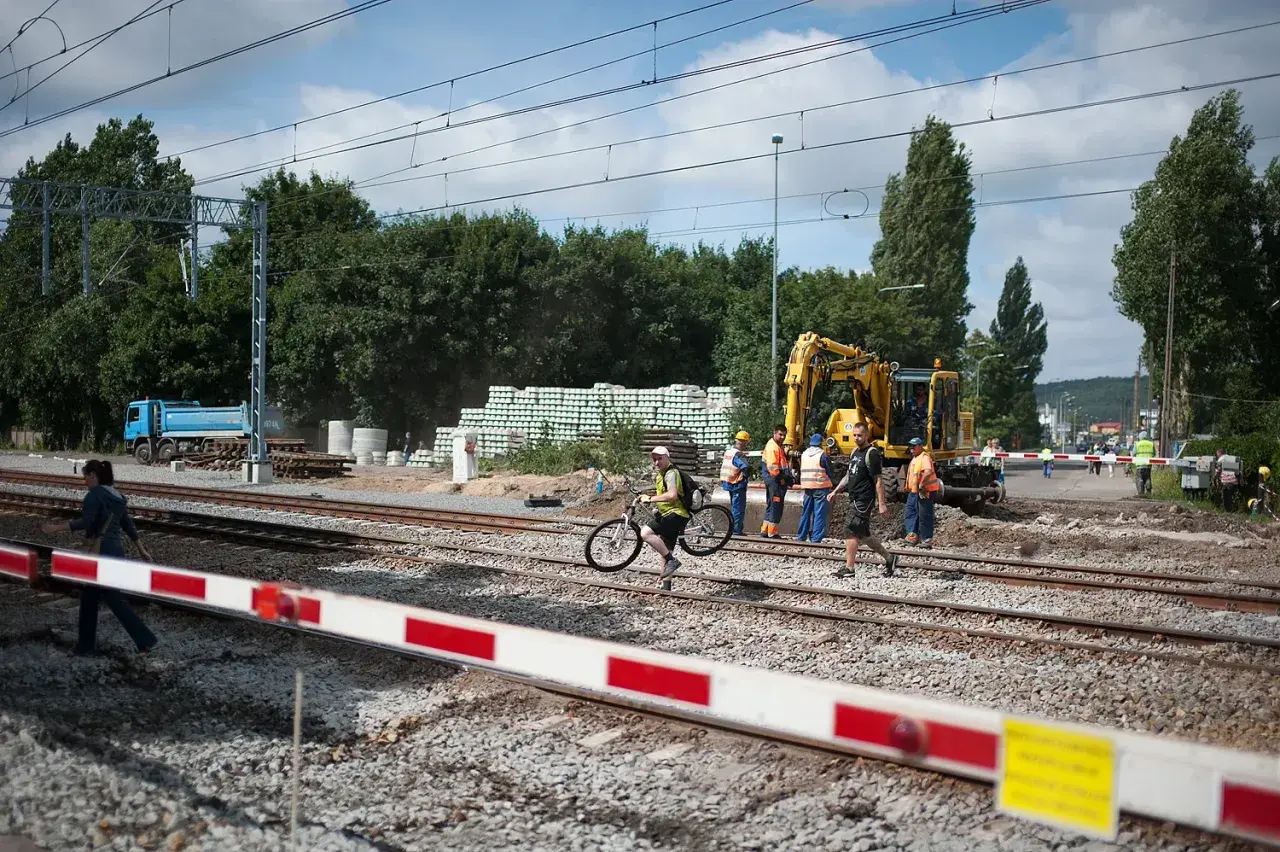 Pracownicy budowlani i dróżnik przejazdowy nadzorują prace przy torach. Koparka i ciężarówka są na miejscu.