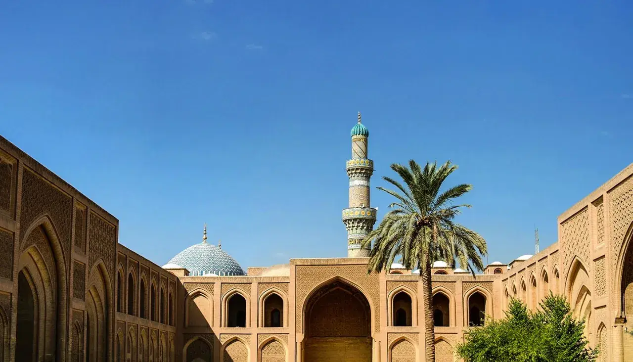 Ein Minarett und eine Kuppel einer Moschee ragen über traditionelle Gebäude mit Bögen und Palmen. Einblick in die reiche irakische Kultur.