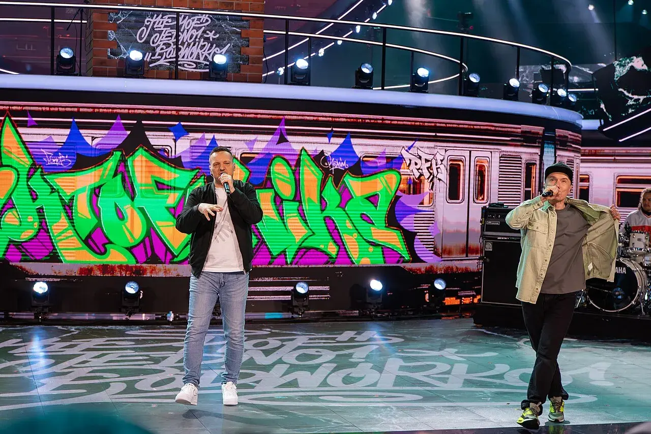Raperzy na scenie, w tle graffiti na wagonie metra. To scena z koncertu, gdzie brzmią **paktofonika utwory**.