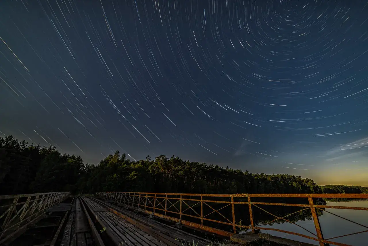 Nocne niebo nad starym mostem, gdzie gwiazdy kreślą na ciemnym tle świetliste ślady.