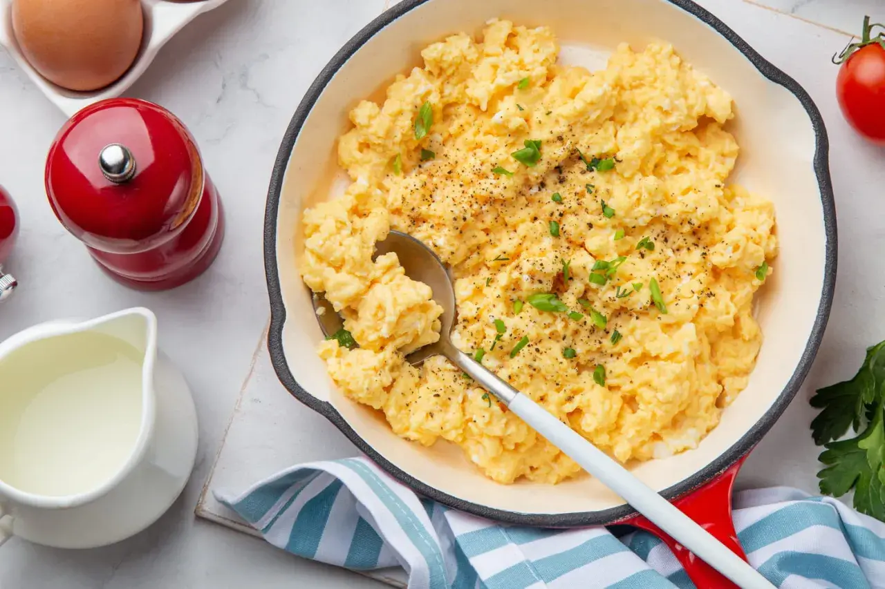 Oeufs brouillés crémeux, parsemés de ciboule fraîche et de poivre noir, servis dans une poêle rouge. Un délice pour le petit-déjeuner.