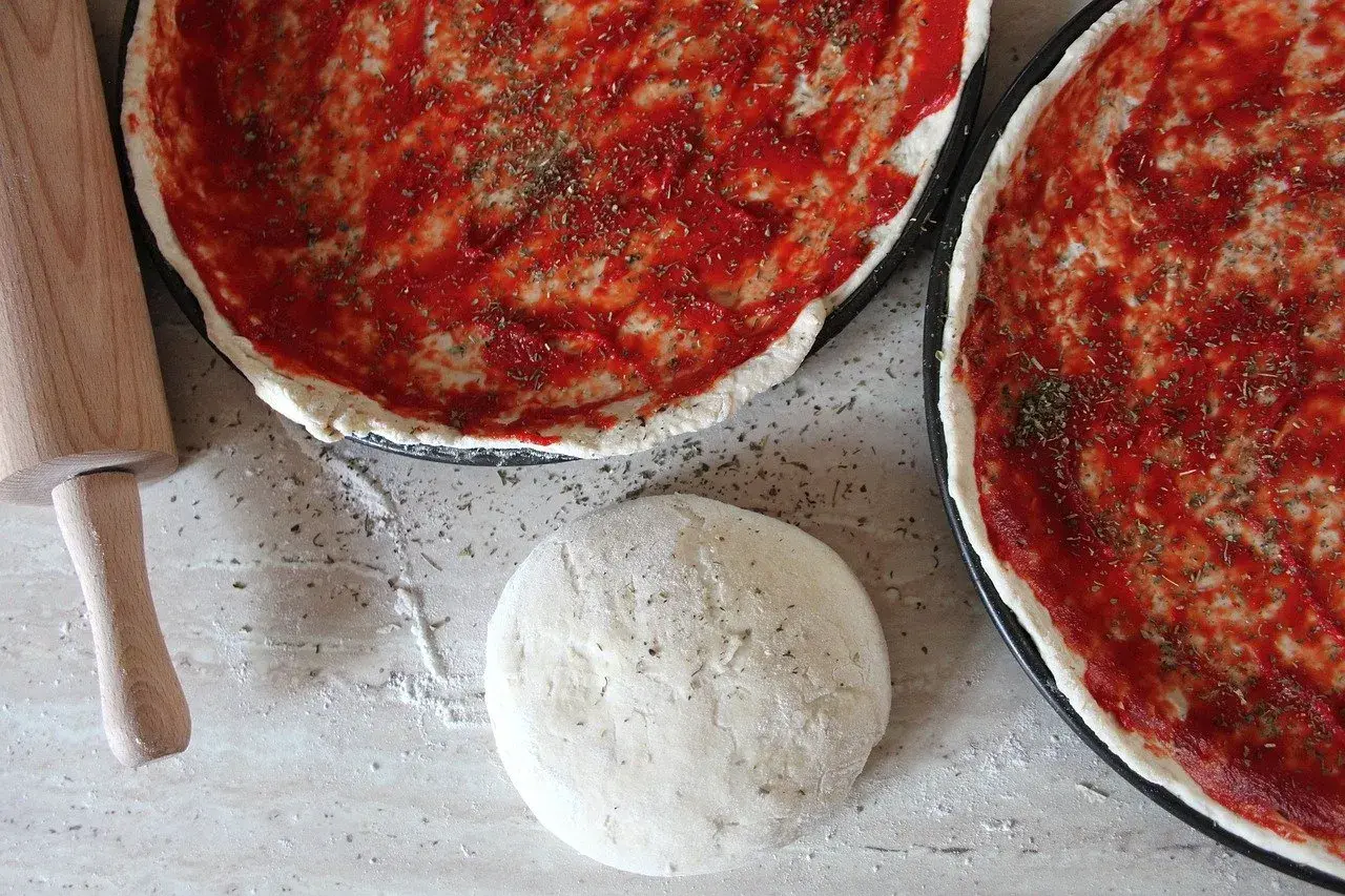 Jak zrobić ciasto na pizzę? Na blacie leży kulka ciasta, wałek i dwie blachy z sosem pomidorowym i ziołami.