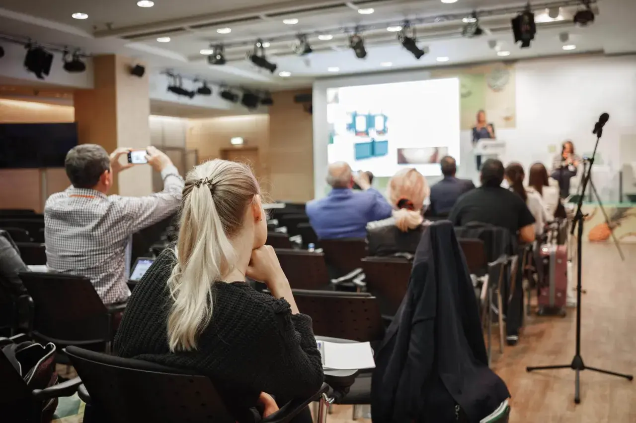 Uczestnicy konferencji, gdzie odbywają się **usługi szkoleniowe**. Jedna osoba filmuje prezentację telefonem.
