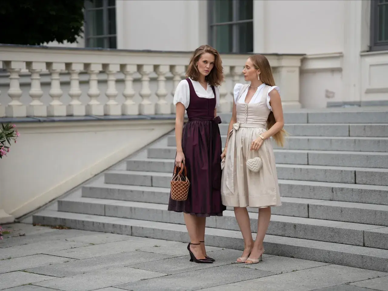 Zwei Frauen in Dirndln vor einer Treppe. Die eine trägt einen dunkelroten Dirndl mit Spitzenbesatz am Rock, die andere einen hellbeigen. Dirndl unterrock ja oder nein? Hier wird die Frage stilvoll beantwortet.