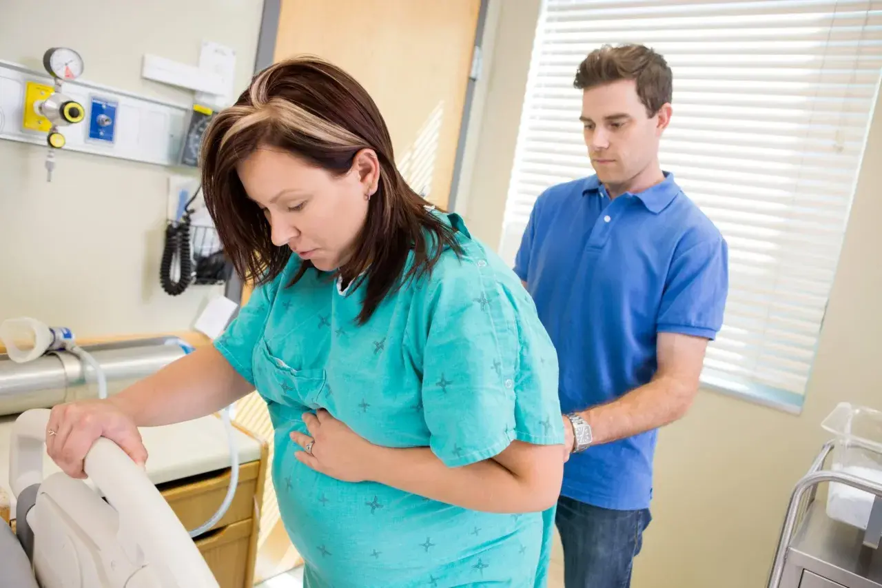 En gravid kvinna håller sig om magen, kanske upplever hon gbs gravid sammandragningar. Hennes partner stöttar henne i sjukhusmiljö.