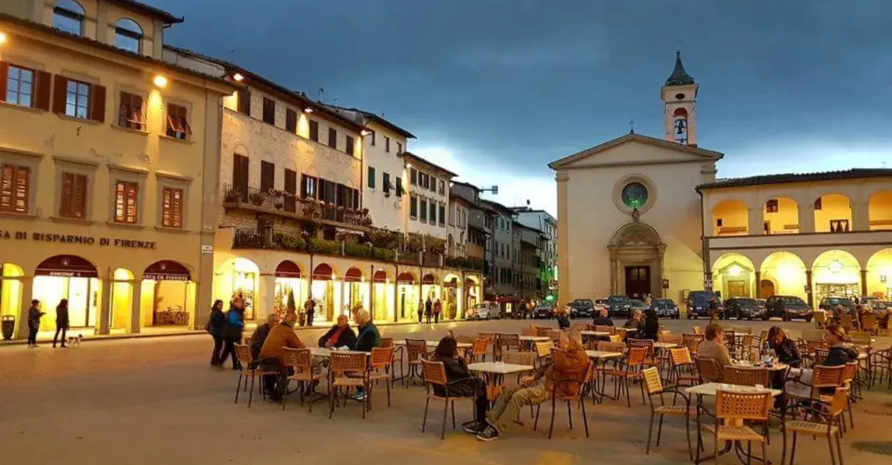 Serata in piazza a Figline Valdarno, con tavolini all'aperto e la chiesa illuminata.