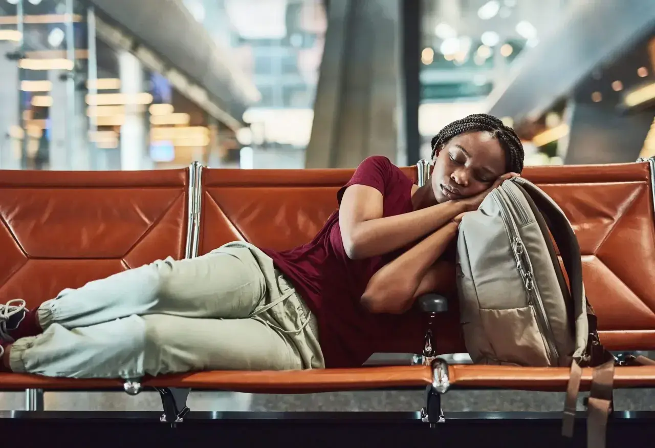 Frau schläft auf einer Bank im Flughafen bei Nacht, ihr Kopf ruht auf ihrem Rucksack.