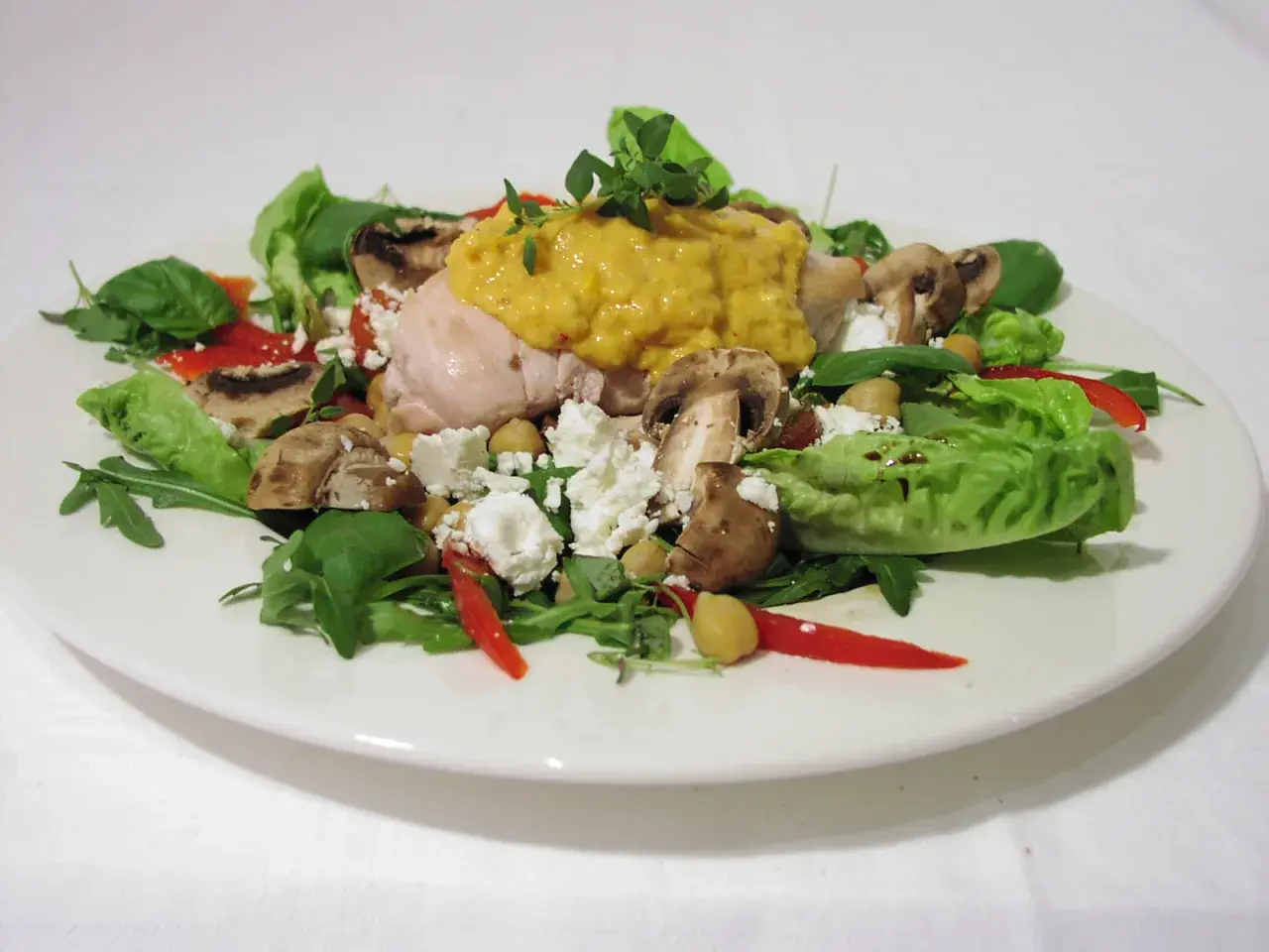 Chicken salad with mushrooms, feta cheese, chickpeas, red pepper, and greens on a white plate.
