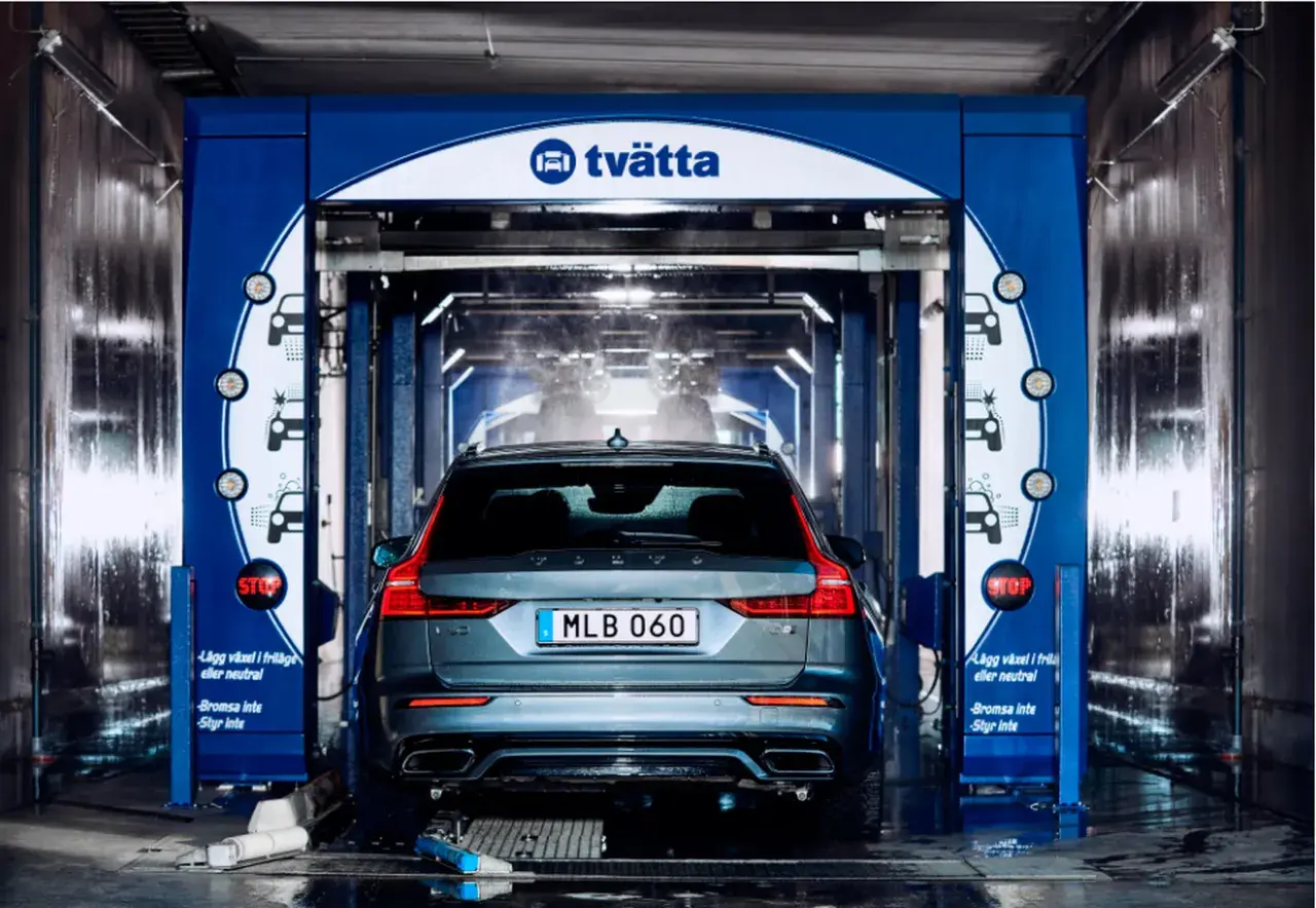 En Volvo är på väg in i en biltvätt. Vatten sprutar och underspolning av bilen pågår.