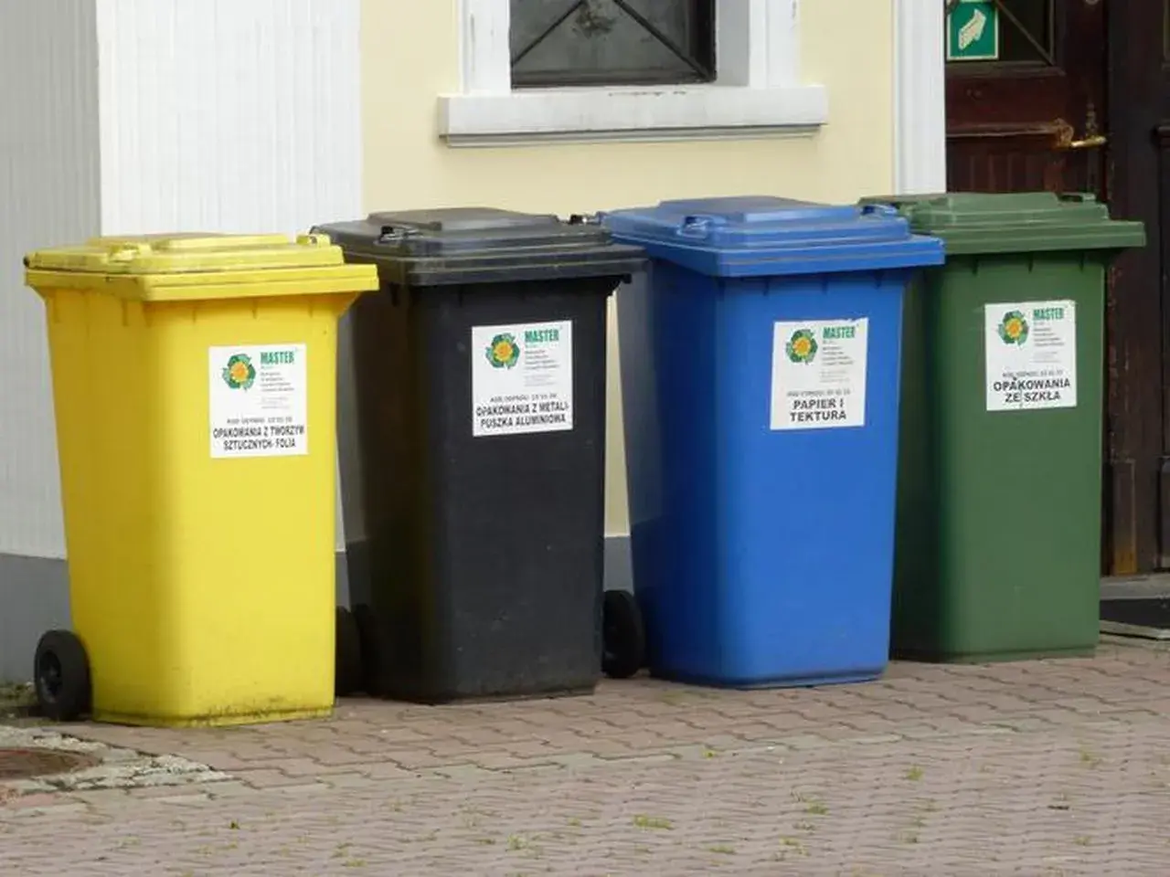 Cztery kolorowe kosze na śmieci: żółty (tworzywa sztuczne), czarny (metale), niebieski (papier) i zielony (szkło). Gdzie wyrzucić opakowanie po galaretce? Do żółtego kosza, jeśli jest plastikowe.