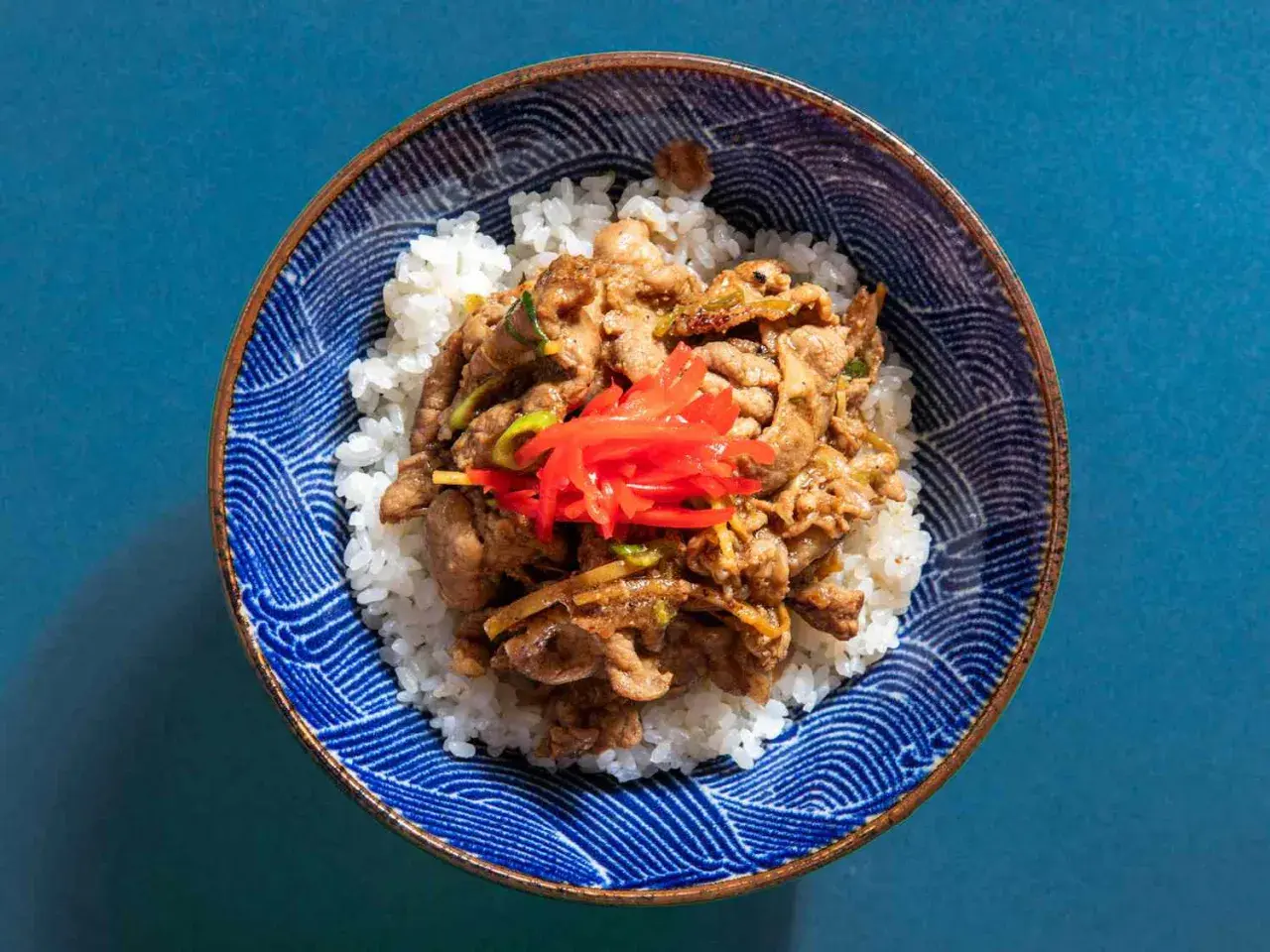 A bowl of fluffy white rice topped with savory pork shogayaki, garnished with bright red pickled ginger.