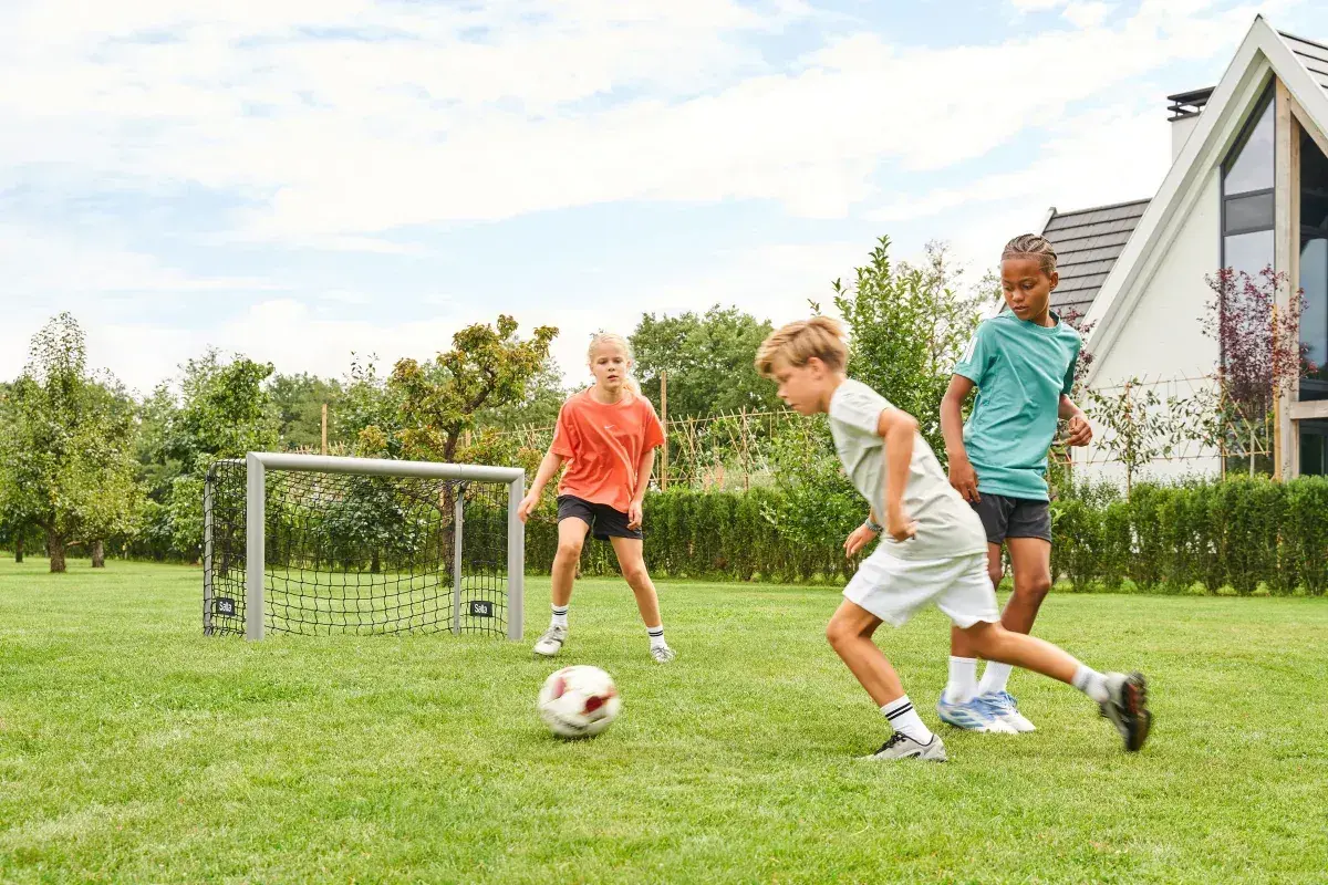 Drei Kinder üben im Garten Fußball. Ein Junge dribbelt, während ein Mädchen und ein weiterer Junge zuschauen.