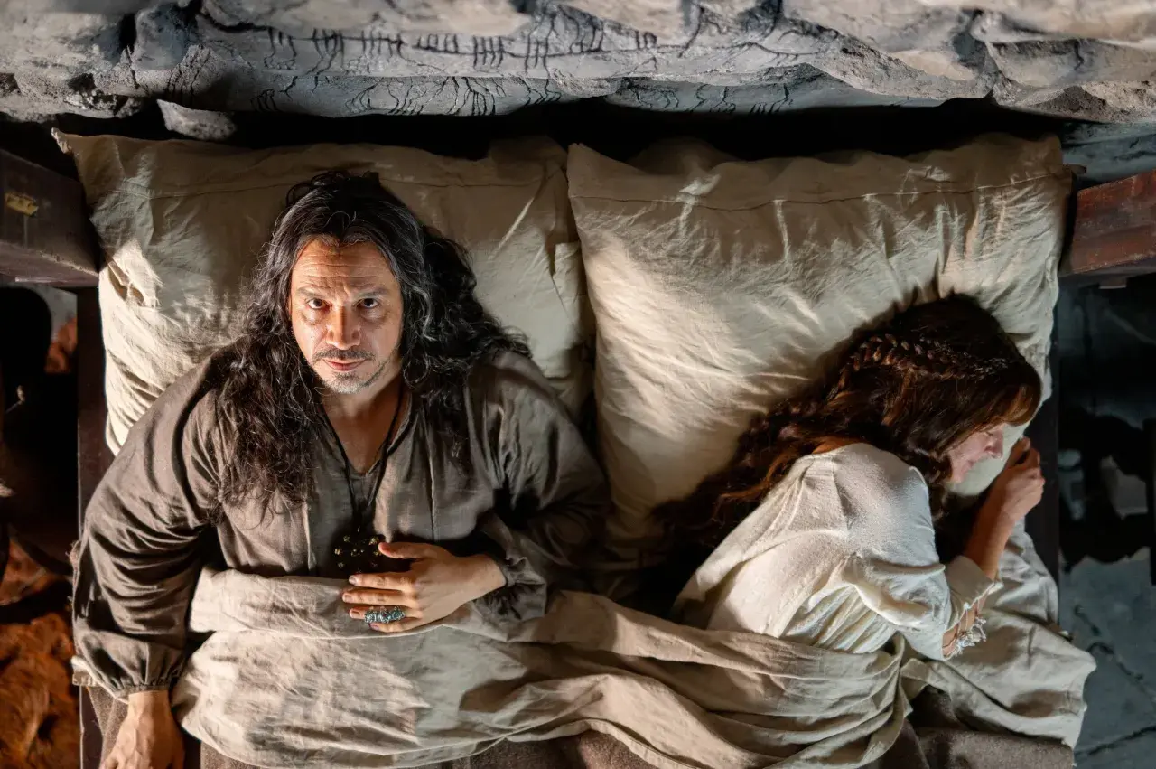 Un homme aux longs cheveux regarde la caméra, allongé dans un lit à côté d'une femme endormie. Un air de mystère plane sur ce moment, rappelant les intrigues de Mordred dans Kaamelott.