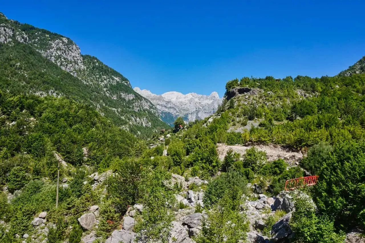 Grüne Hänge und felsige Gipfel in den albanischen Alpen unter strahlend blauem Himmel. Ein kleiner roter Steg führt über einen steinigen Bachlauf.