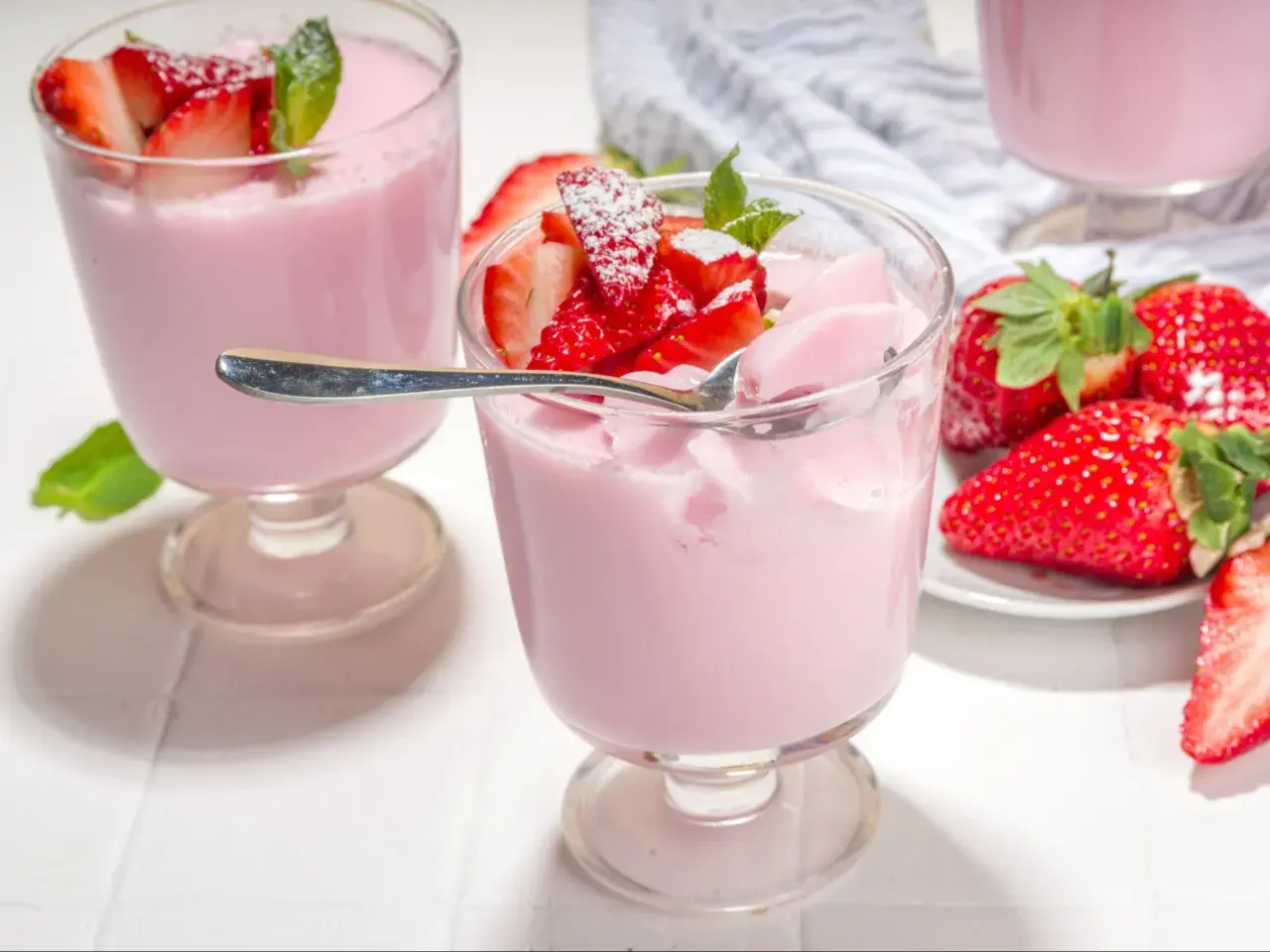 Różowa panna cotta z truskawkami, udekorowana listkami mięty i posypana cukrem pudrem. Obok świeże truskawki.