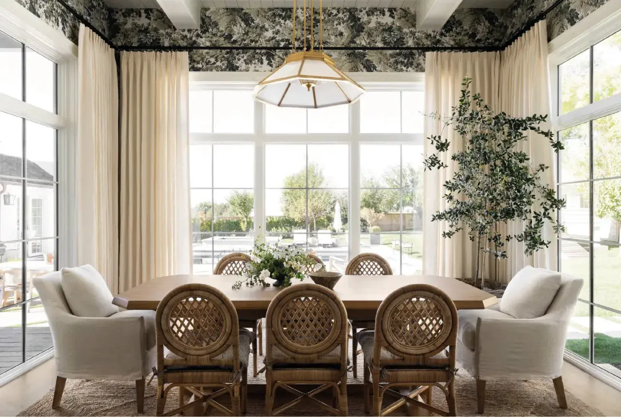 A bright dining room in a Studio McGee home features a large wooden table with rattan chairs, plush armchairs, and a floral wallpaper.