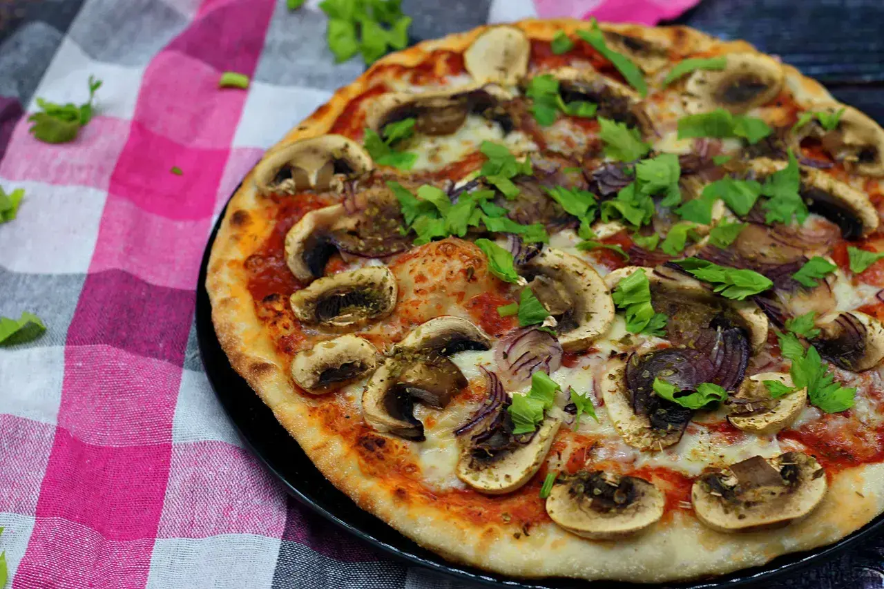 Pyszna pizza z pieczarkami na pizze, cebulą i świeżą natką pietruszki, podana na kraciastej serwetce.