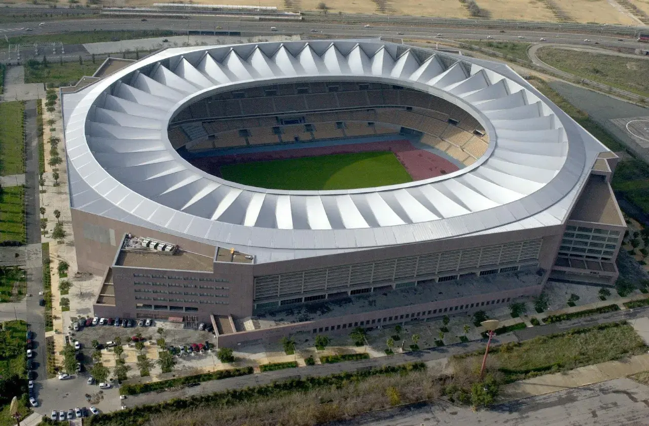 Imponujące, okrągłe **Estadio La Cartuja** z charakterystycznym, falującym dachem, otoczone zielenią i drogami.
