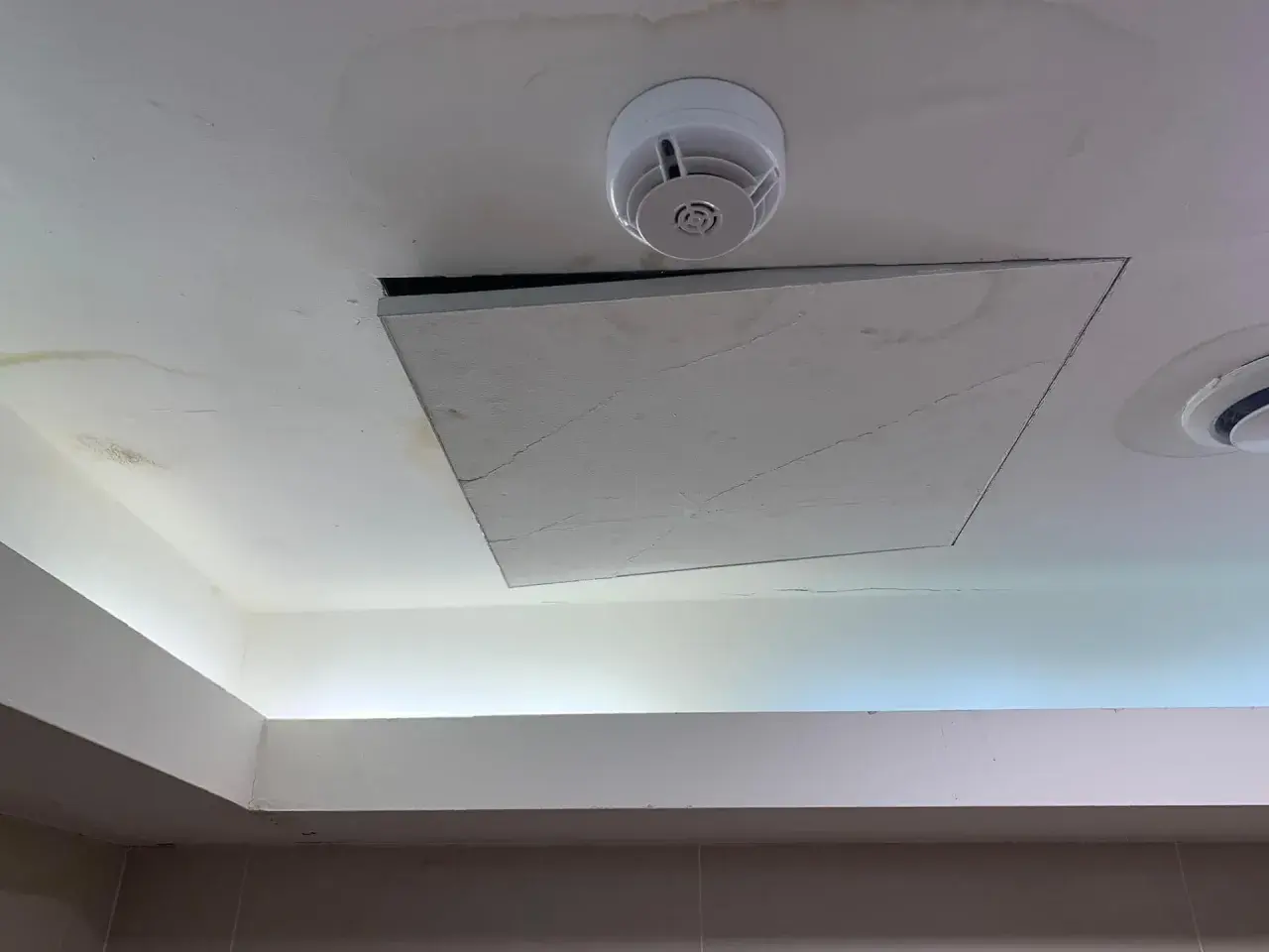 Un soffitto in cartongesso con una botola aperta, un rilevatore di fumo e macchie d'acqua. Il montaggio botola cartongesso è visibile.