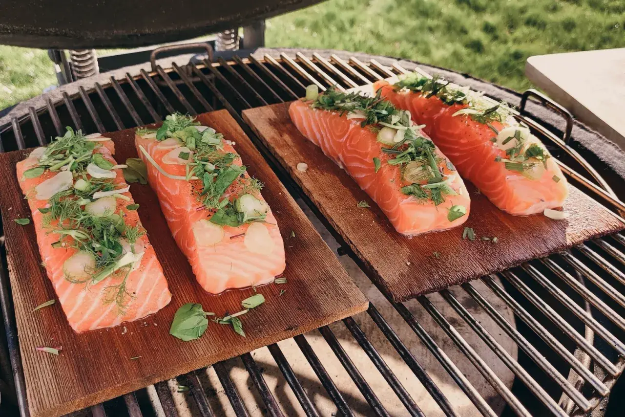 Quatre pavés de saumon sur planche de cèdre, garnis d'herbes fraîches et d'ail, prêts à être grillés.