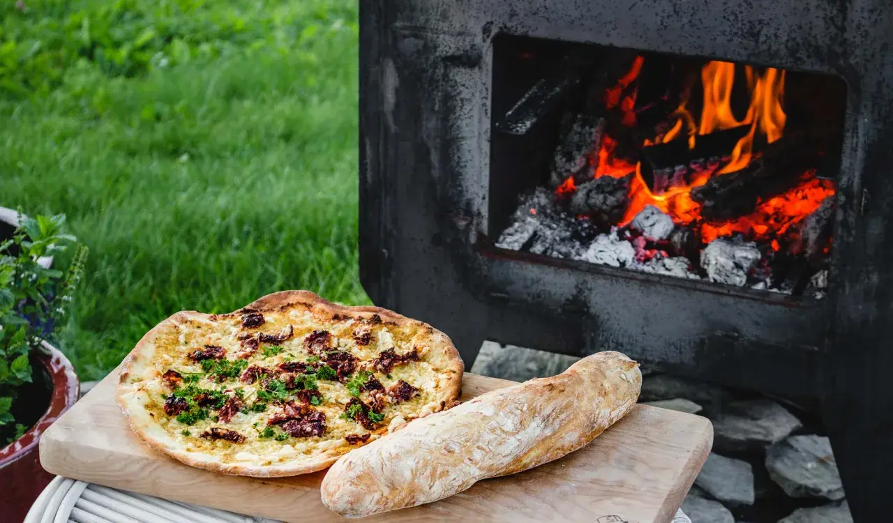 En pizza och ett bröd ligger på en skärbräda bredvid en pizzaugn med glödande ved.