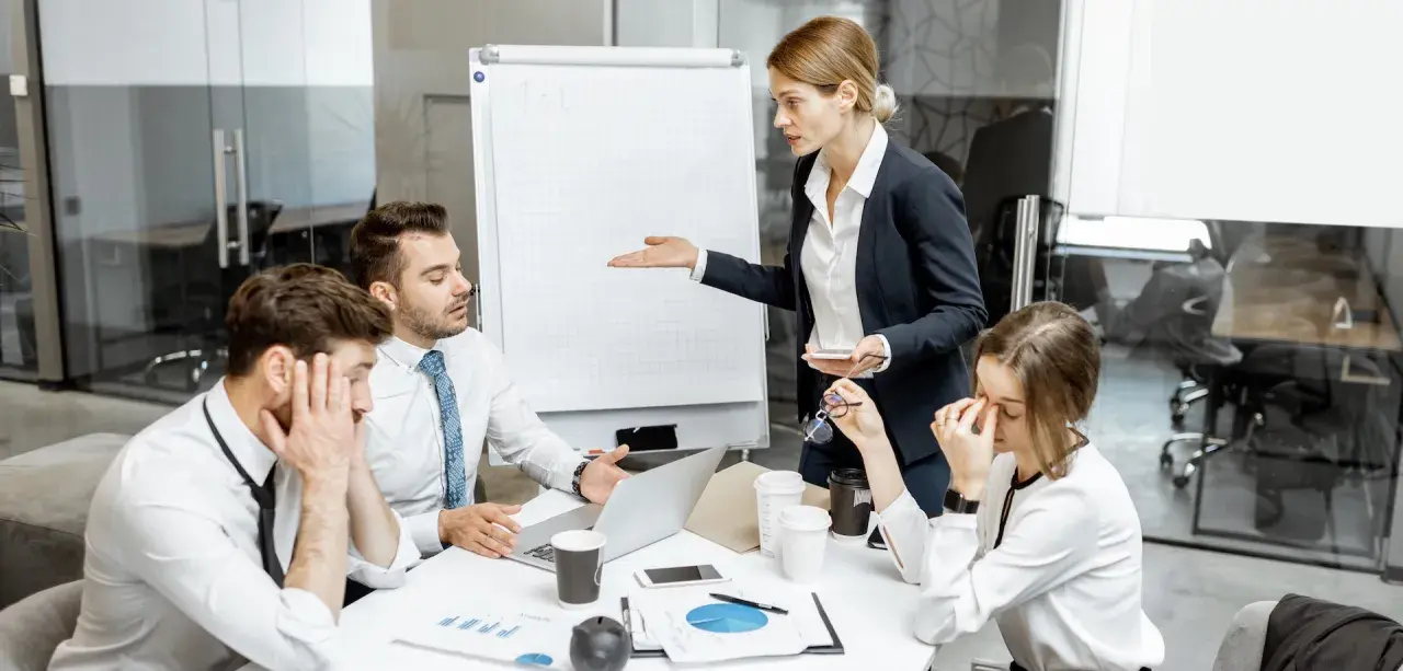 En kvinna gestikulerar mot en whiteboard medan kollegor ser utmattade ut. Känslan av manipulativt beteende hänger i luften under mötet.